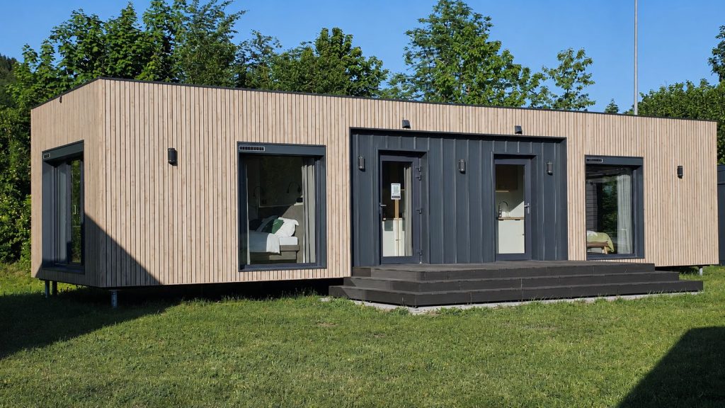 Alpenchalet Modulhaus mit heller Holzfassade und anthrazitfarbenem Eingang auf sonniger Wiese, Lieferung in die Steiermark.
