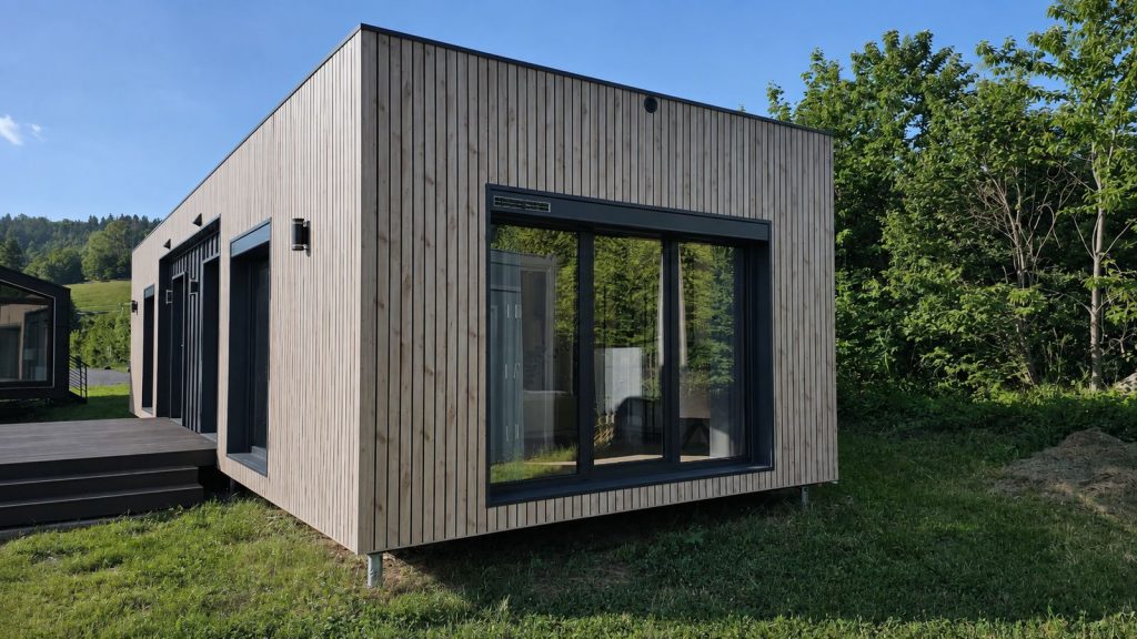 Alpenchalet mit heller Holzfassade und bodentiefer Schiebetür, Premium-Modulhaus von BOND vor bewaldeten Hügeln.