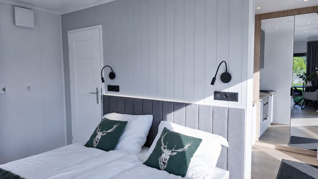 Helles Schlafzimmer im Alpenchalet mit grauem Polsterbett, Hirschmotiv-Kissen und offenem Durchblick zur Kueche - Modulhaus von BOND.