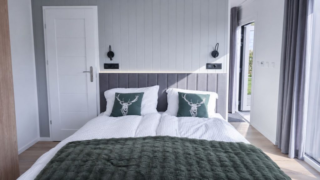 Schlafzimmer mit grau gepolstertem Kopfteil, Hirsch-Kissen und grüner Tagesdecke - schlüsselfertig ausgestattet.