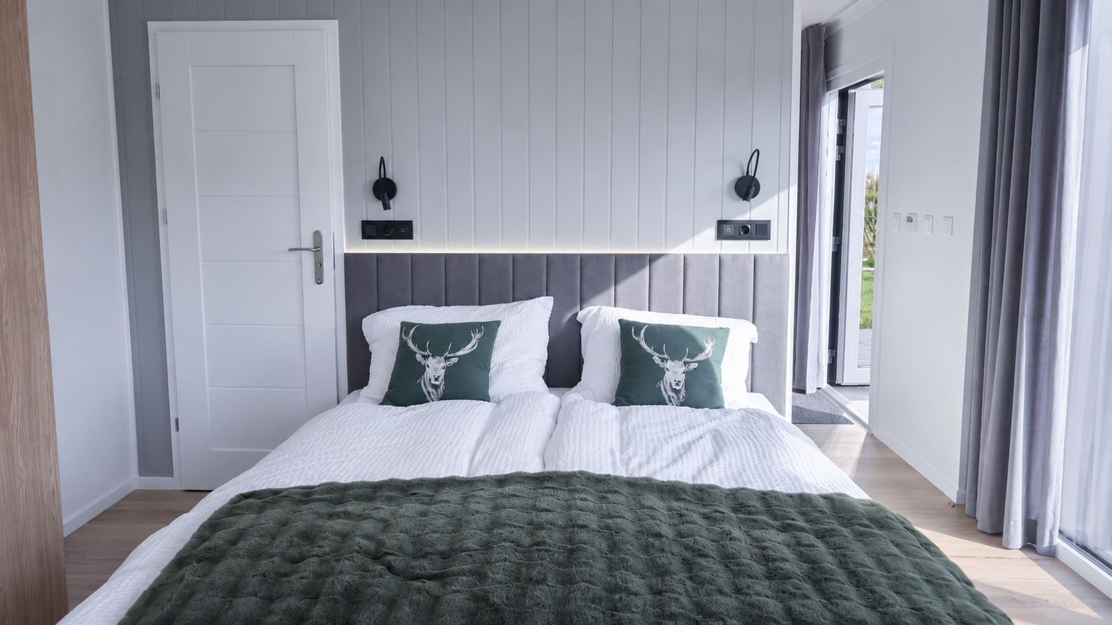 Schlafzimmer mit grau gepolstertem Kopfteil, Hirsch-Kissen und grüner Tagesdecke - schlüsselfertig ausgestattet.