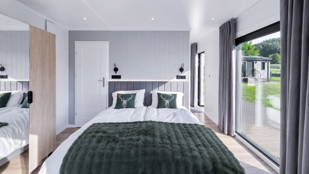 Helles Schlafzimmer mit Doppelbett, gruener Tagesdecke und Hirsch-Kissen an bodentiefer Glasfront - wohnliches Ganzjahreshaus.