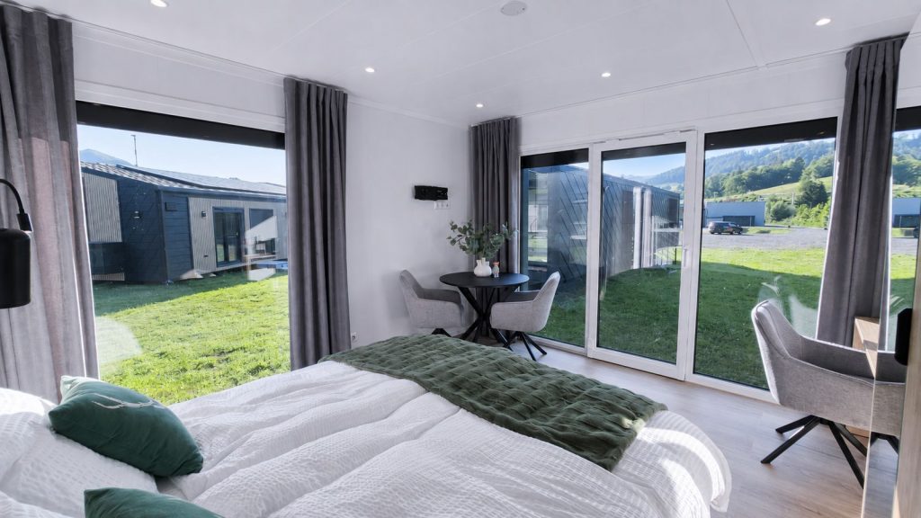 Helles Schlafzimmer mit bodentiefen Fenstern, grünen Textilakzenten und kleiner Sitzecke - wohnfertiges Ganzjahreshaus von BOND.