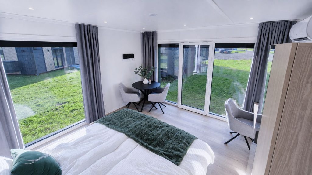 Helles Schlafzimmer mit bodentiefen Fenstern, gruenem Plaid und Sitzecke im Modulhaus - Lieferung nach Salzburg.