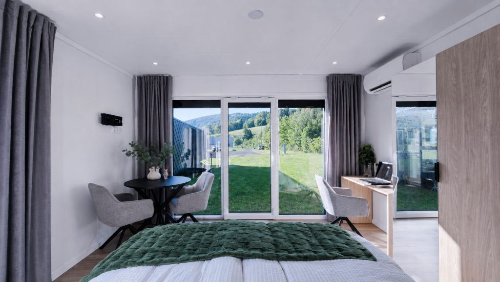 Helles, vollmoebliertes Schlafzimmer mit bodentiefen Fenstern, gruenem Ueberwurf und kleiner Sitzecke am Panoramafenster.