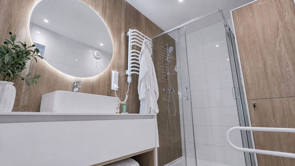 Badezimmer mit rundem LED-Spiegel, Holzoptik-Wand, Aufsatzwaschbecken und ebenerdiger Glasdusche im Fertighaus.