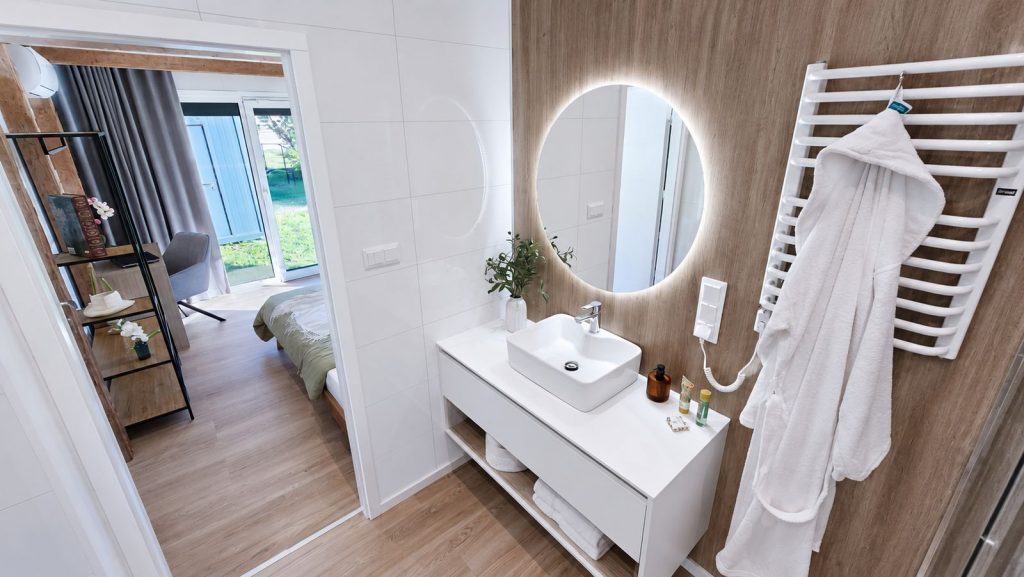 Badezimmer mit rundem LED-Spiegel, Holz-Akzentwand und Aufsatzwaschbecken, Durchblick ins Schlafzimmer - wohnfertig uebergeben.