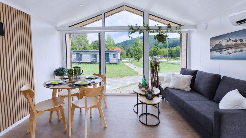 Wohn-Essbereich mit raumhoher Giebelverglasung, Holzlamellenwand und rundem Esstisch - lichtdurchflutetes Premium-Mobilheim.