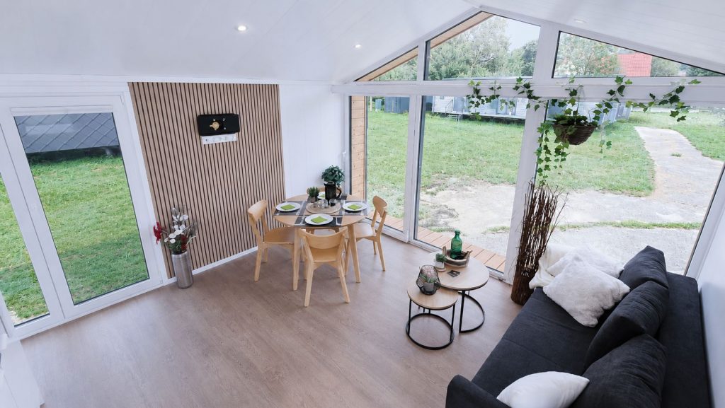 Fertigteilhaus mit offenem Wohn-Essbereich, Giebelverglasung, runder Essgruppe und dunklem Sofa mit Gartenblick.