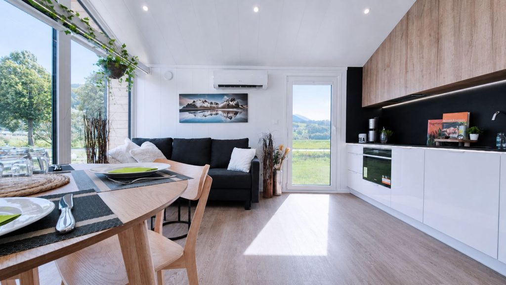 Wohnkueche mit dunklem Sofa, Holz-Esstisch, weisser Kuechenzeile und Wandbild mit Bergmotiv - schluesselfertig eingerichtet.