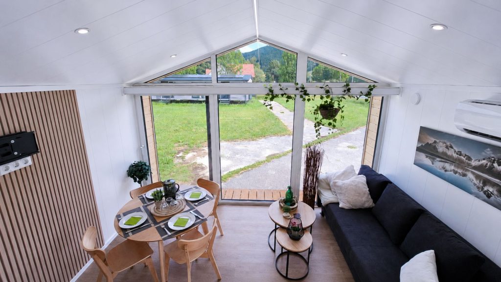 Wohnbereich mit Giebel-Panoramafenster, Essgruppe und dunklem Sofa - Ganzjahreshaus, verfuegbar in Villach.