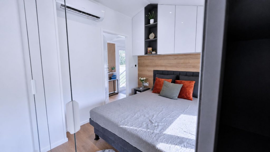 Schlafzimmer mit weissem Schrank, Holzakzent und orangen Kissen in durchdachter Massivmodul-Architektur.