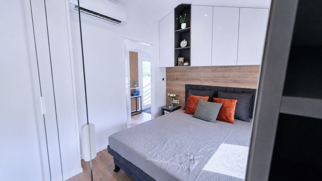 Schlafzimmer im Diamond Family Deluxe mit Holzkopfwand, orangen Akzentkissen und Klimageraet - mobiles Eigenheim mit viel Stauraum.
