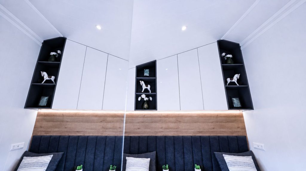 Detail des Bettkopfteils im Premium-Modulhaus: Holzpaneel mit LED-Licht, Samt-Polsterung und offene Deko-Nischen.