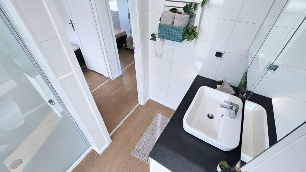 Blick ins Badezimmer mit Aufsatzwaschbecken auf schwarzer Platte, Dusche und weissen Fliesen - vollmoebliert uebergeben.
