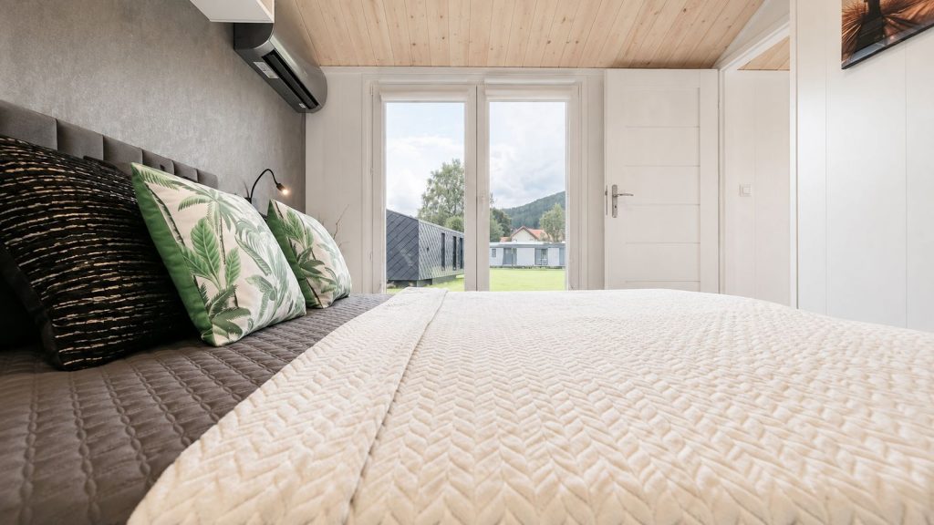 Wohnfertiges Schlafzimmer mit cremefarbener Tagesdecke, Palmenkissen und bodentiefer Gartentuer.