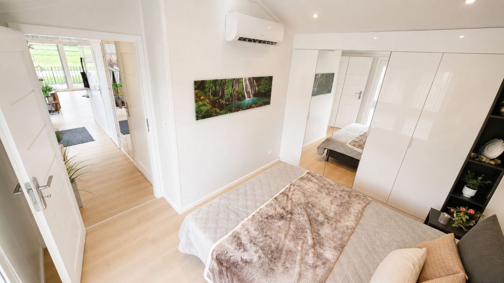 Vollmoebliertes Schlafzimmer mit Klimageraet, Hochglanz-Spiegelschrank und Blick in den offenen Wohnbereich.