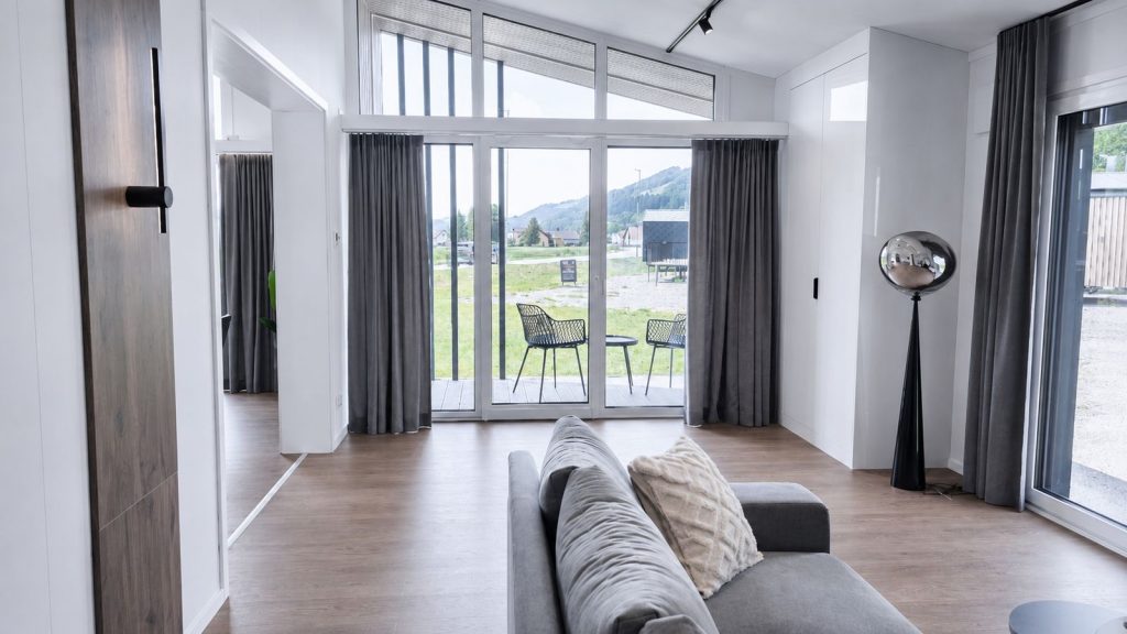 Wohnzimmer mit grauem Sofa, bodentiefer Giebelverglasung und Terrassenzugang im modernen Eigenheim.
