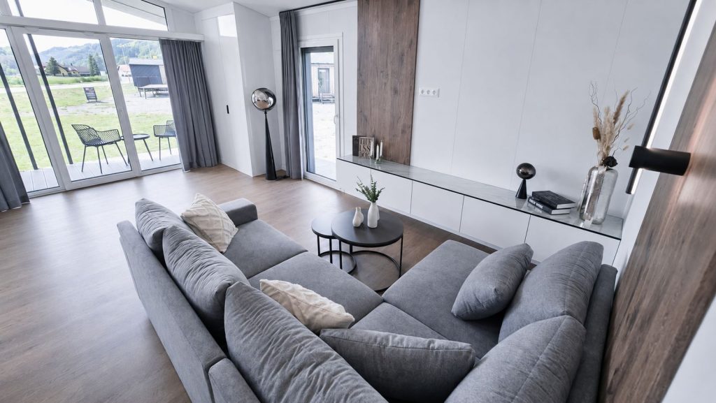Wohnzimmer mit grauer Ecksofalandschaft, runden Beistelltischen und Panoramafenstern, modernes Eigenheim mit Terrassenzugang.