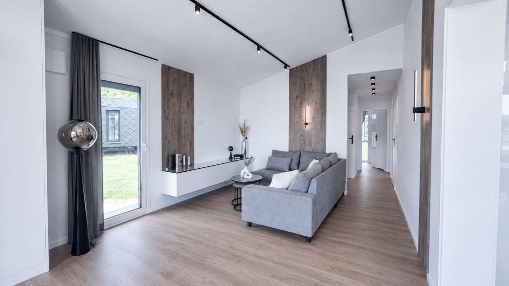 Schluesselfertiger Wohnraum mit grauem Sofa, Holzakzentwand, Stehleuchte und langem Flur mit Schienenlicht.