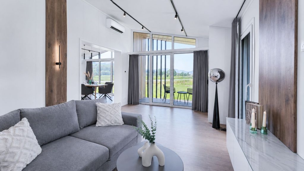 Wohnzimmer mit grauem Sofa, Bogenleuchte und Glasschiebetuer zur Terrasse mit Wiesenblick - elegantes Premium-Modulhaus.