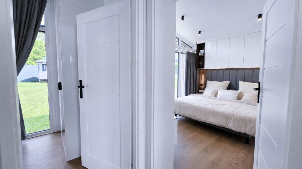 Blick durch weiße Türen ins Schlafzimmer mit Doppelbett im Diamond Vario Deluxe, Fertighaus für Klagenfurt.