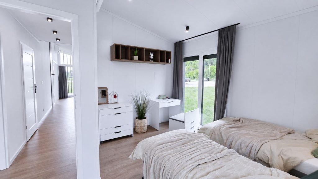 Schlafzimmer mit zwei Betten, weissem Schreibtisch, Kommode und bodentiefer Terrassentuer im Premium-Modulhaus.