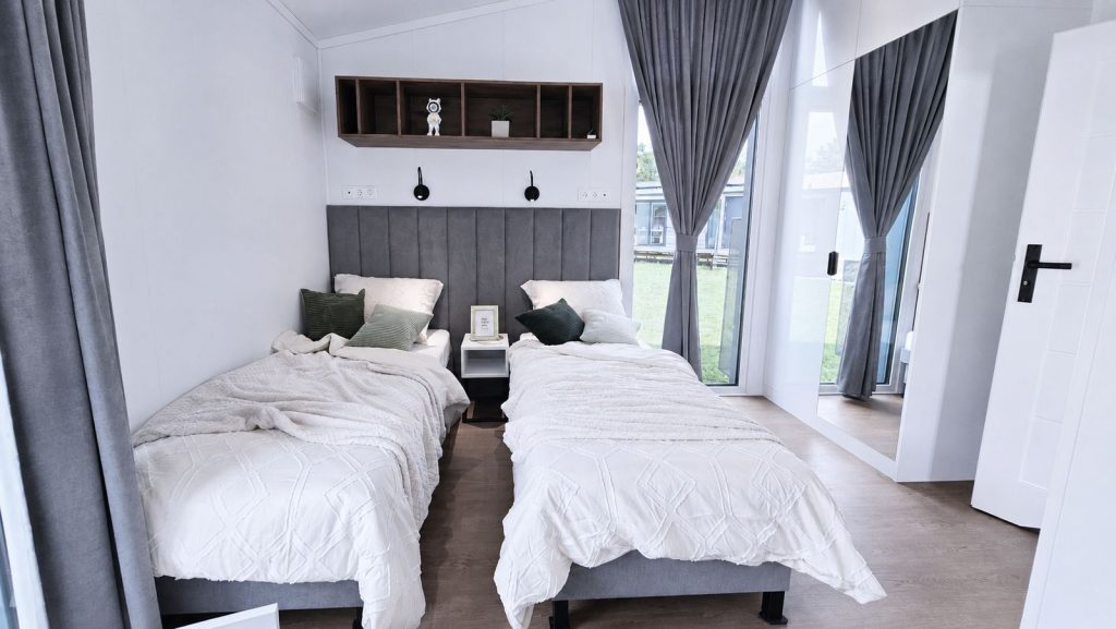 Wohnfertiges Zweibettzimmer mit gepolsterter Kopfteilwand, Regalbord und Schiebetuer zum gepflegten Gartengrund.