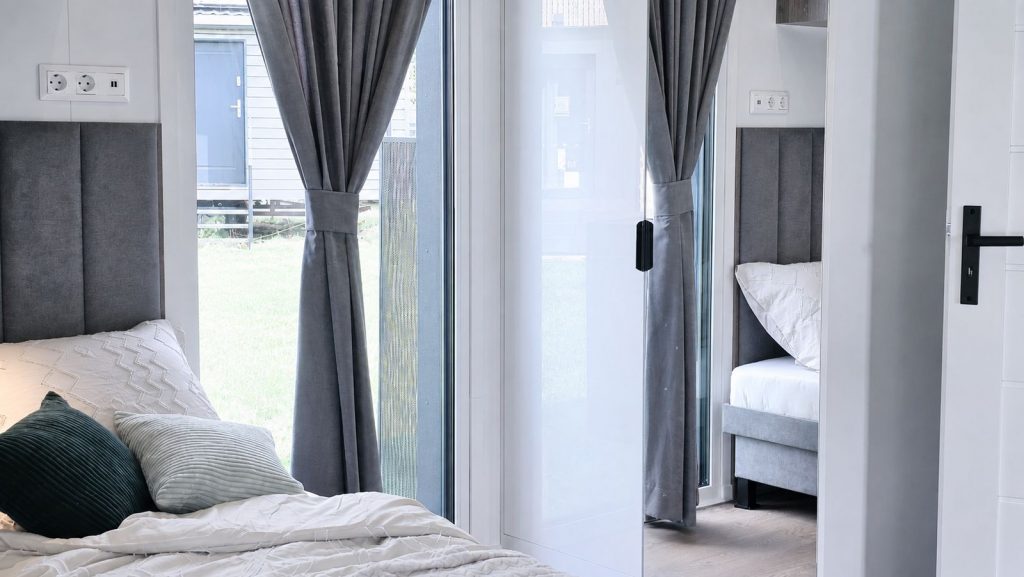 Schlafbereich mit bodentiefem Fenster, grauen Vorhaengen und Blick ins zweite Schlafzimmer, mobiles Eigenheim von BOND.