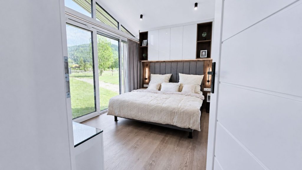 Schlafzimmer im Diamond Vario Deluxe mit Doppelbett, Oberschränken und großer Fensterfront - Massivmodul-Architektur.