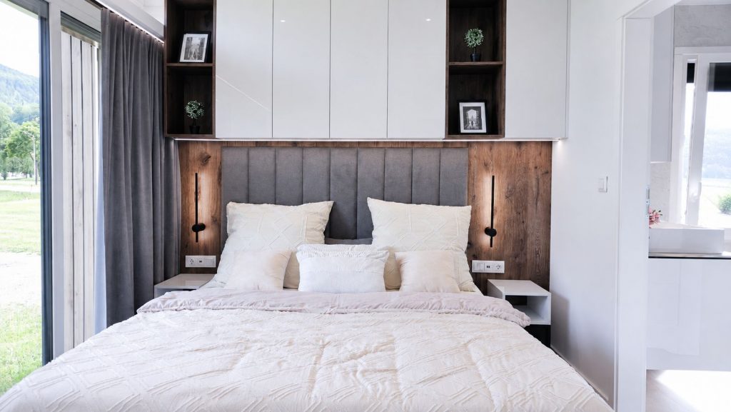 Schlafzimmer im Diamond Vario Deluxe mit Holz-Akzentwand, grauem Kopfteil und cremefarbener Bettwaesche - mobiles Eigenheim.