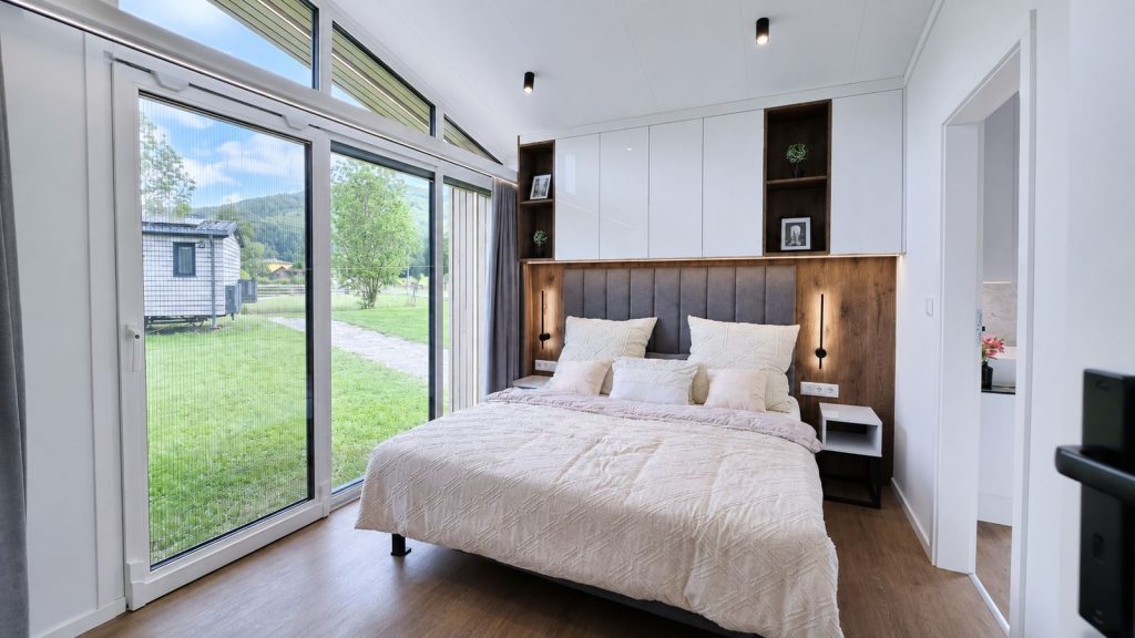 Schlafzimmer mit Holz-Kopfteil, indirektem Licht und bodentiefer Verglasung zum Garten, Premium-Mobilheim fuer Tirol.