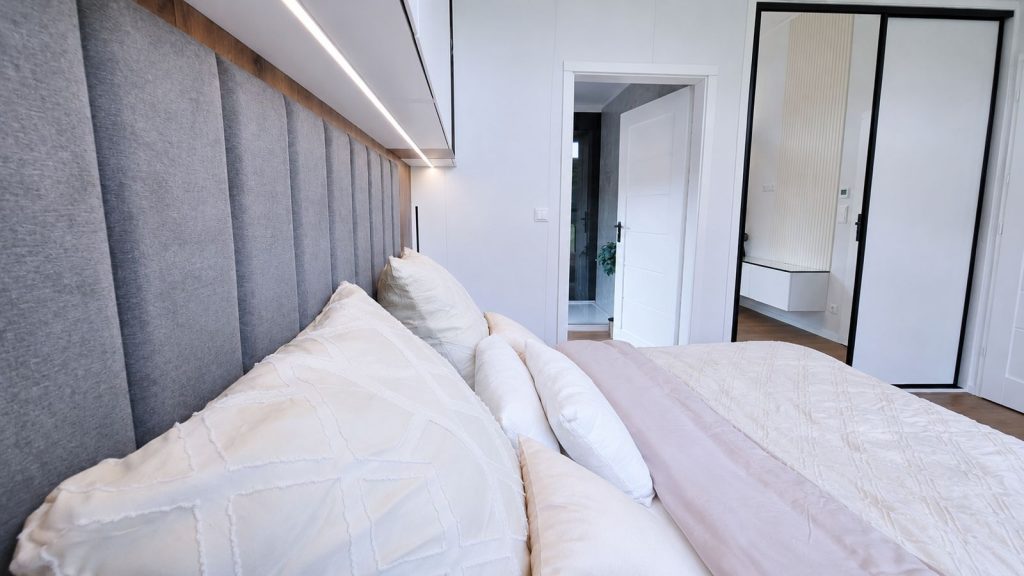 Schlafzimmerdetail mit gepolstertem Kopfteil, indirekter Beleuchtung und Spiegelschrank im Premium-Modulhaus.