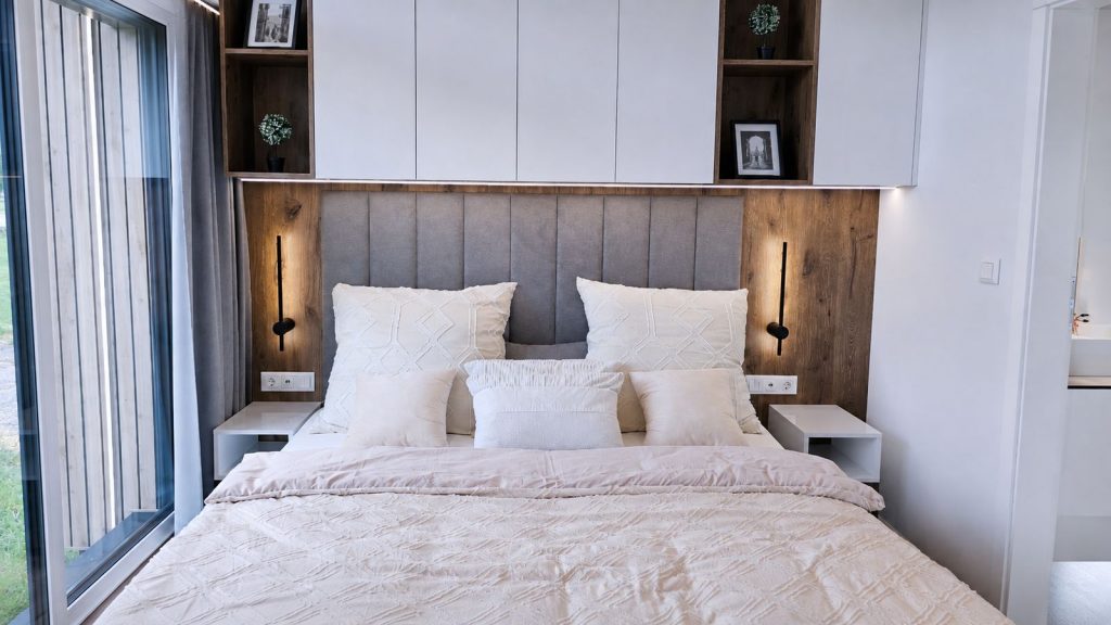 Schlafzimmer mit grauem Polsterkopfteil zwischen Holzpaneelen, cremefarbener Bettwaesche und Oberschraenken - Fertigteilhaus fuer Graz.