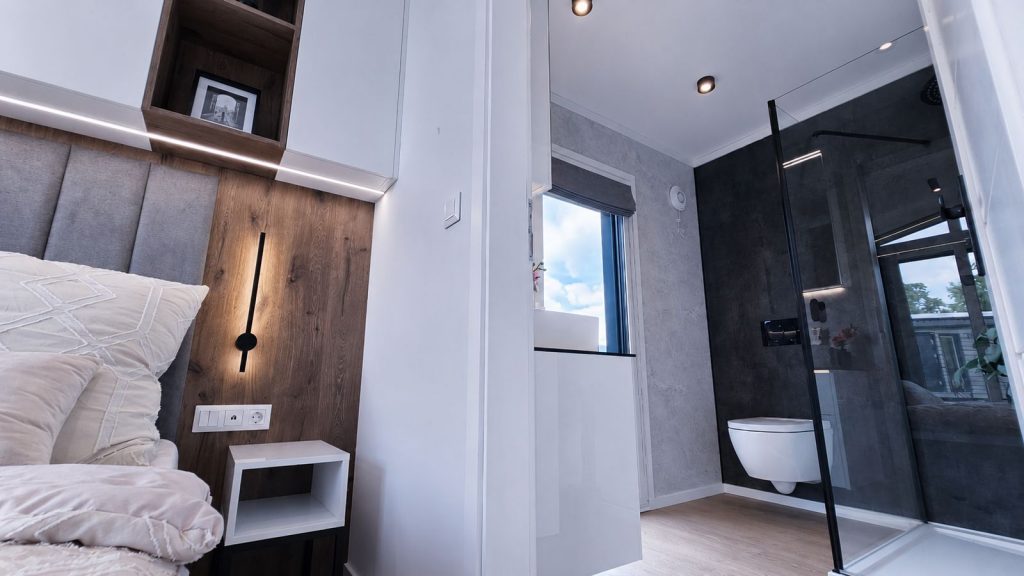 Schlafbereich mit Holz-Akzentwand neben Duschbad mit Wand-WC in Betonoptik - Ganzjahreshaus, verfuegbar in Wels.