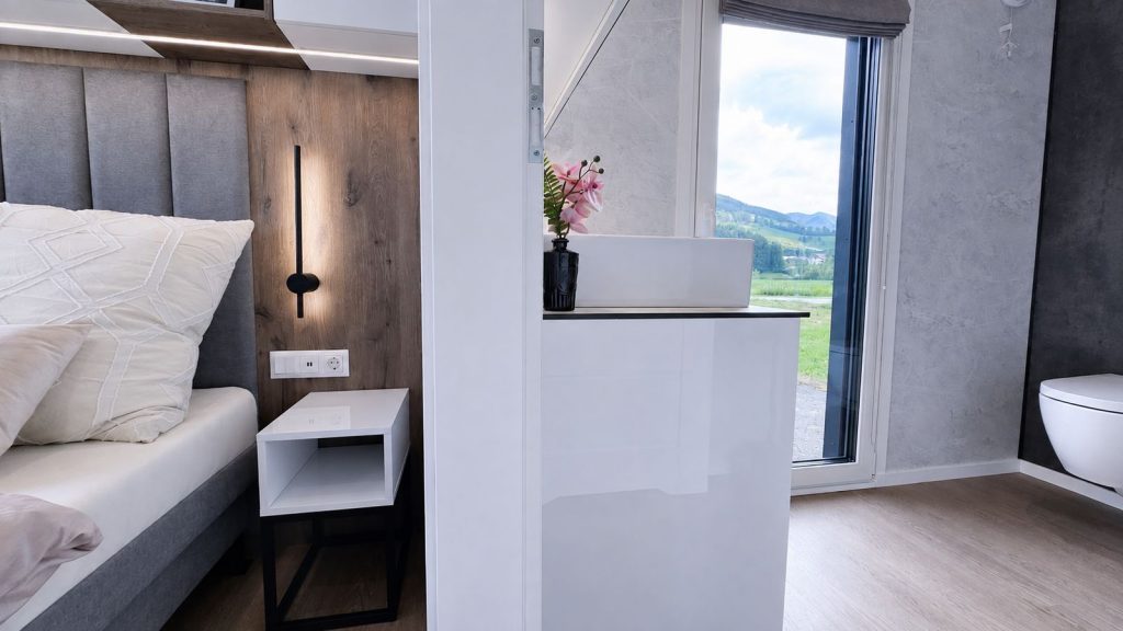 Schluesselfertiges Schlafzimmer mit Holzwand, Wandleuchte und angrenzendem Waschbereich mit Orchidee.