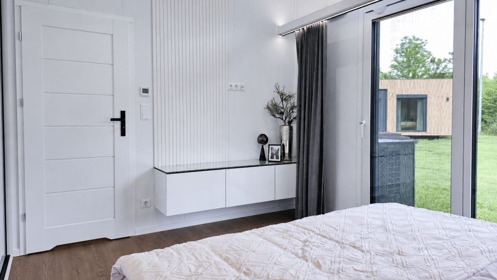 Schlafzimmer mit weisser Lamellenwand, schwebendem Sideboard und Schiebetuer zur Terrasse - Modulhaus von BOND.