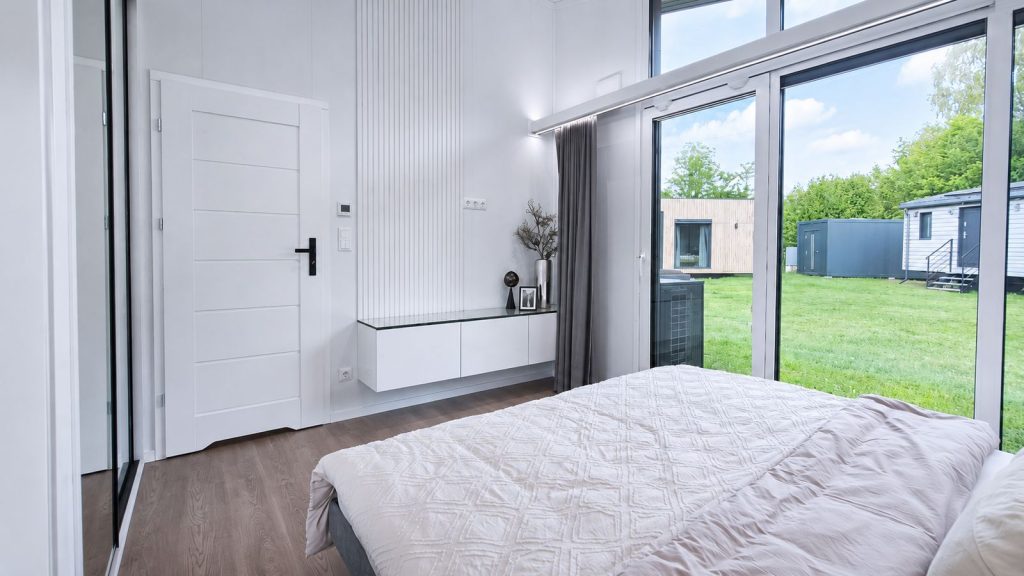 Schlüsselfertiges Schlafzimmer mit bodentiefer Fensterfront, schwebendem Sideboard und Blick in die grüne Umgebung.