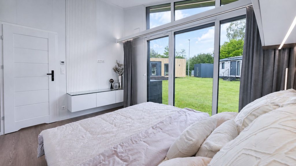 Schlafzimmer mit Doppelbett, bodentiefer Verglasung und Sideboard mit Glasplatte im Premium-Modulhaus - verfuegbar im Burgenland.
