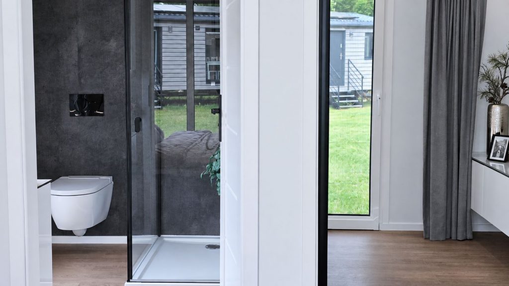 Ensuite-Bad mit ebenerdiger Dusche und Wand-WC vor dunkler Steinoptik, praezise Massivmodul-Architektur mit Gartenblick.
