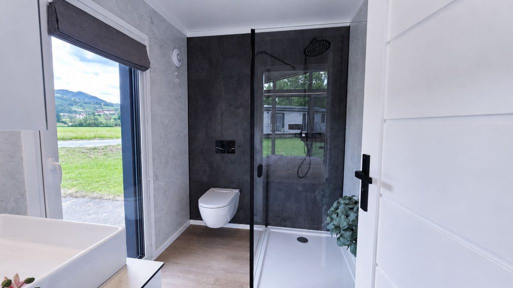 Bad in Steinoptik mit Regendusche, schwarz gerahmtem Glas und Fensterblick ins Gruene - Premium-Modulhaus, verfuegbar in Kaernten.