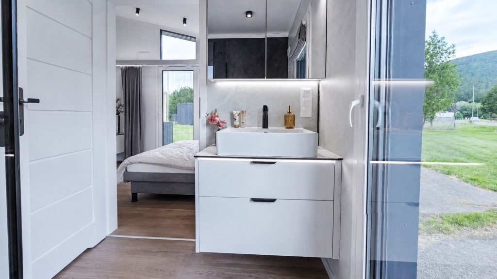 Bad en suite mit weissem Aufsatzwaschbecken und Marmoroptik-Wand, Durchblick zum Schlafzimmer - Modulhaus fuer das Burgenland.