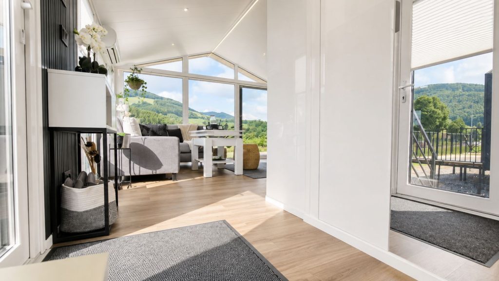 Wohnzimmer im Modulhaus Diamond XS Deluxe mit Giebelfenster, grauem Sofa und Bergblick - verfügbar in Villach.
