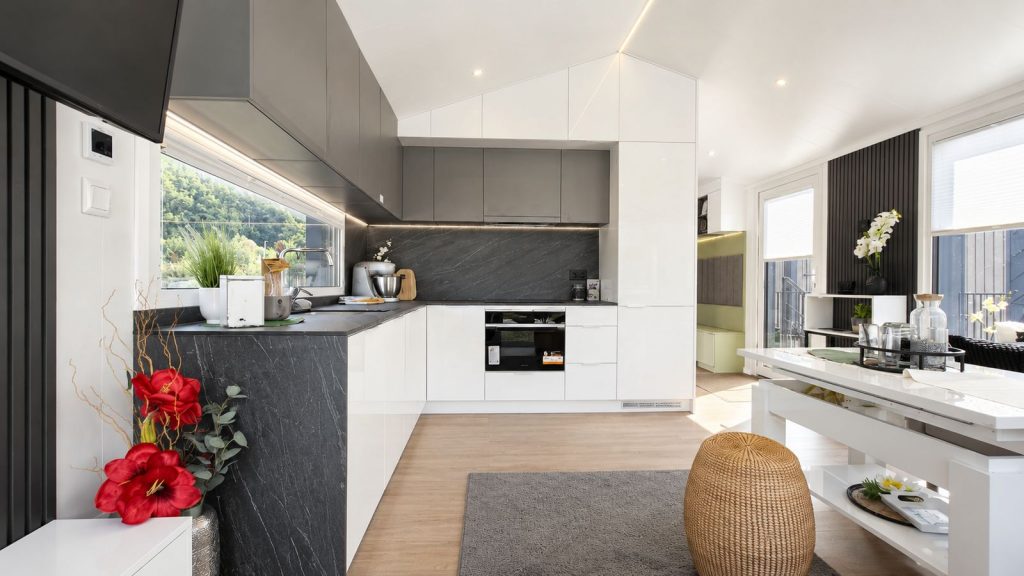 L-Kueche im Diamond XS Deluxe mit weissen Hochglanzfronten, dunkler Steinoptik-Rueckwand und Backofen - Premium-Modulhaus fuer Innsbruck.