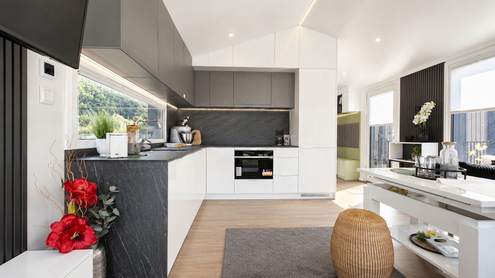 L-Kueche im Diamond XS Deluxe mit weissen Hochglanzfronten, dunkler Steinoptik-Rueckwand und Backofen - Premium-Modulhaus fuer Innsbruck.