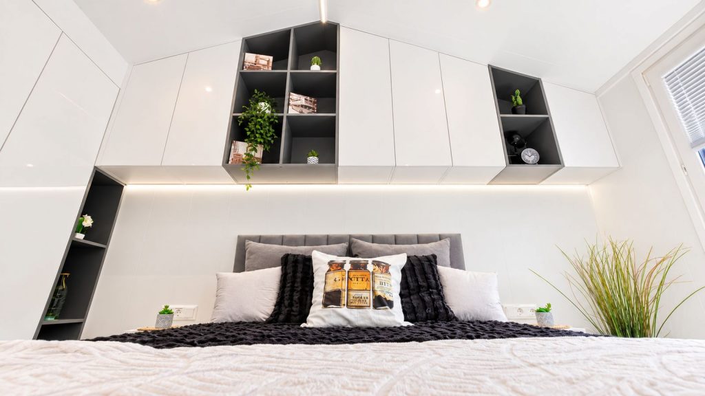 Vollmoebliertes Schlafzimmer mit weissen Schraenken, offenen Wuerfelregalen und gepolstertem Bettkopf.
