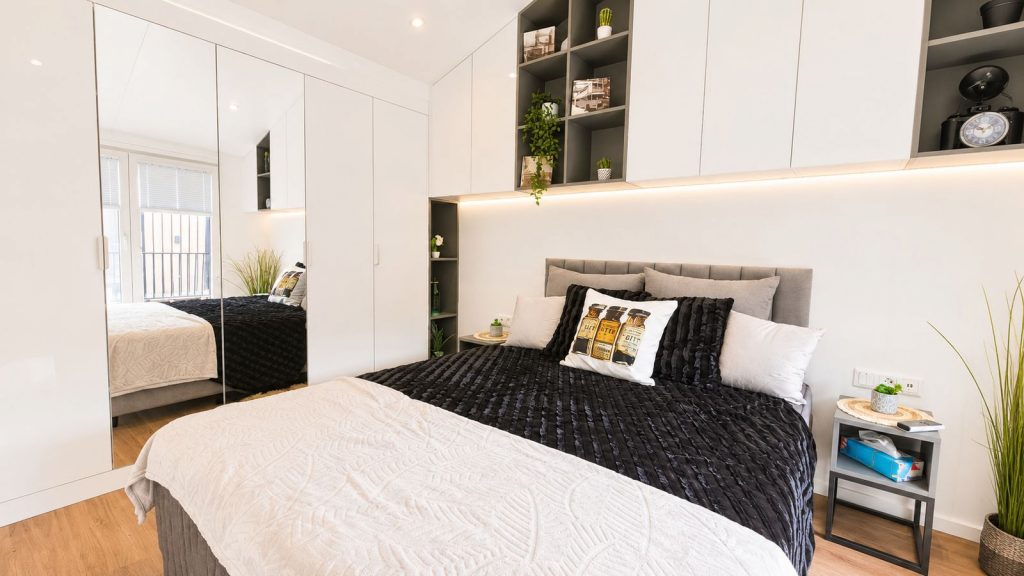 Schlafzimmer mit Spiegelschrank, Regalnischen und dunkler Tagesdecke im Premium-Mobilheim mit indirekter LED-Beleuchtung.