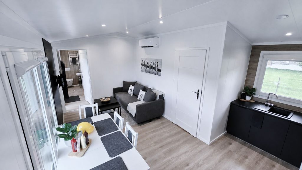 Vollmöblierter Wohnraum mit grauem Sofa, weißem Esstisch, schwarzer Küchenzeile und Blick ins Badezimmer.