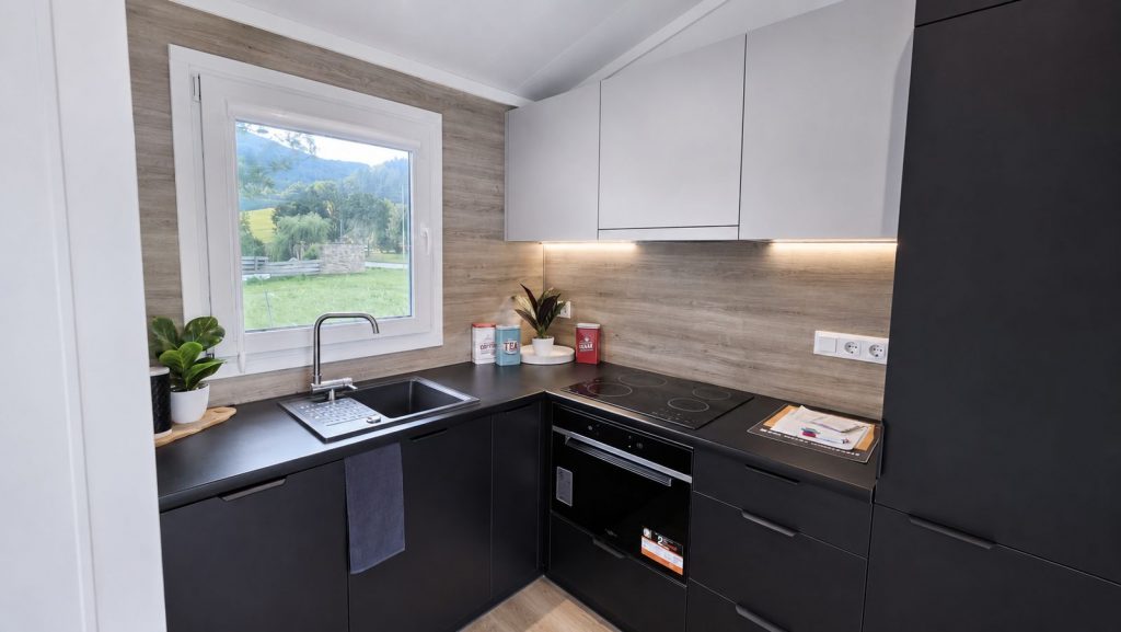 Kompakte Kueche mit schwarzen Fronten, Holzoptik-Rueckwand und Fenster zur Wiese - vollmoebliert geliefert.