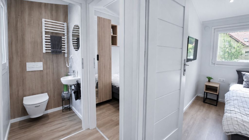 Blick vom Bad mit Holzwand und Wand-WC ins Schlafzimmer mit TV, durchdachte Raumaufteilung im Premium-Mobilheim.